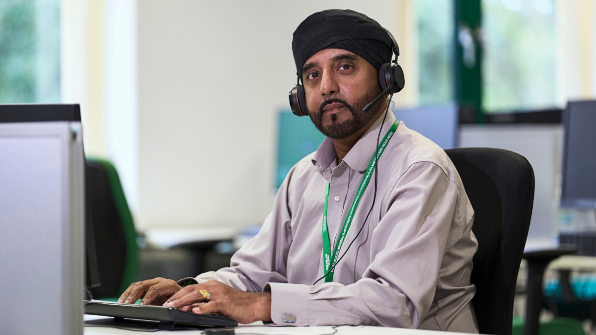 A photo of Akbal, a person who works on the Macmillan Support Line. Akbal is seated at a desk and typing into a computer keyboard, while wearing a phone headset.