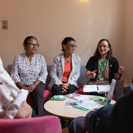 A group of people are sitting around a coffee table. Nisha (pictured right) is sitting on the end of a sofa. She has a notebook open in her lap. She is wearing a dark cardigan and colourful top. 
