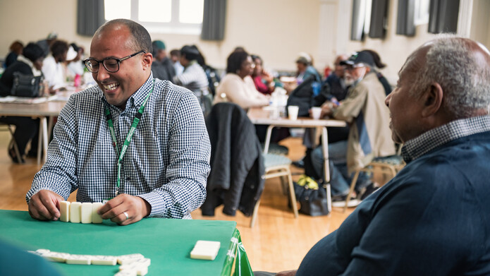 Two men are playing dominoes on a table inside a large conference room. Behind them are other tables with people playing dominoes and chatting. They are all members of the Pineapple Club in South East London.