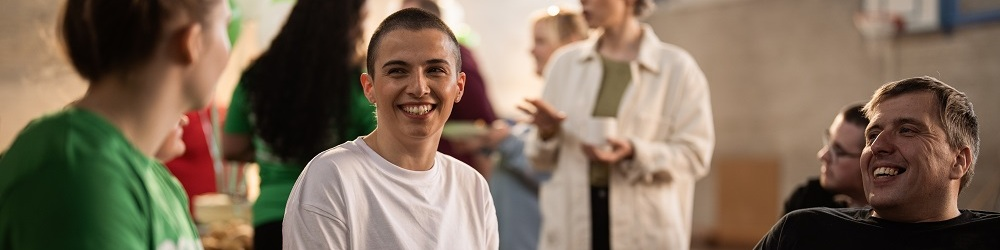 Three people are sitting and chatting at a Coffee Morning. A person with short hair and wearing a white tshirt is holding a plastic drink cup. Next to them is a person in a black tshirt holding a plate with food and a plastic drink up. Behind them are other people at the event. They appear to be in an outdoor basketball court. 