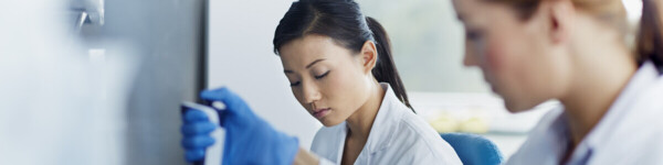 Two female scientists working in a laboratory. One is using a pipette to handle a sample, while the other writes notes on a document. 