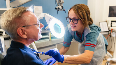 A Macmillan speech and language therapist is with a patient in a hospital consulting room. She is examining the patient's neck. 