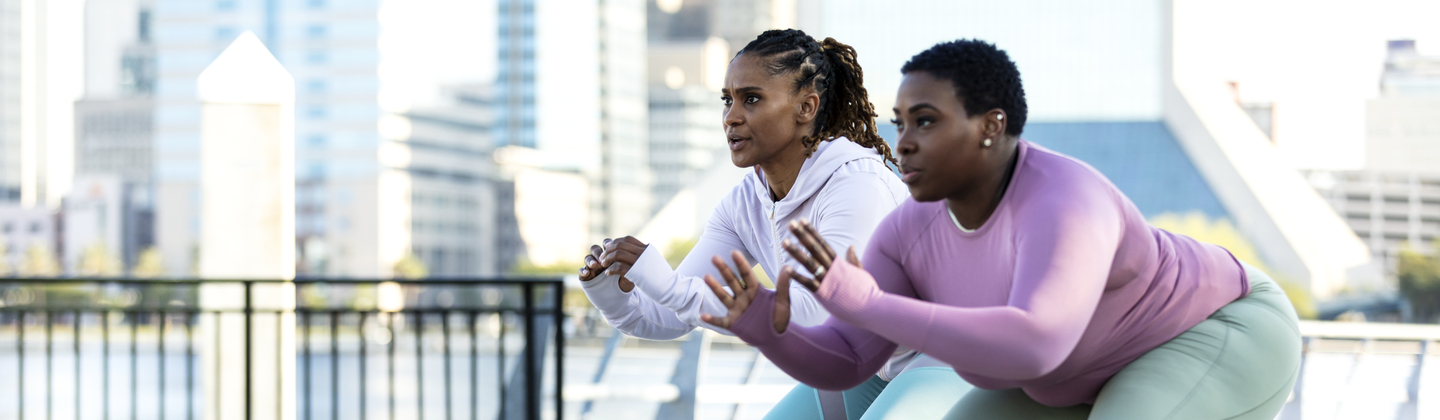 Two people are exercising outside. They are using resistance bands and are in a squat position. They are wearing sports clothing. 