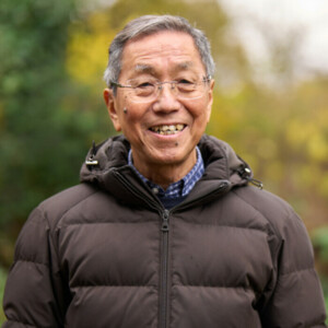 Man outdoors, smiling while wearing a dark padded jacket. The background shows greenery. 