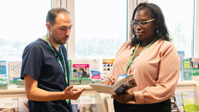 Two professionals review a booklet; one in scrubs explains, while the other, in a pink blouse, listens. Shelves of healthcare brochures fill the background.