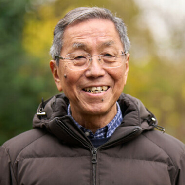 Man outdoors, smiling while wearing a dark padded jacket. The background shows greenery. 