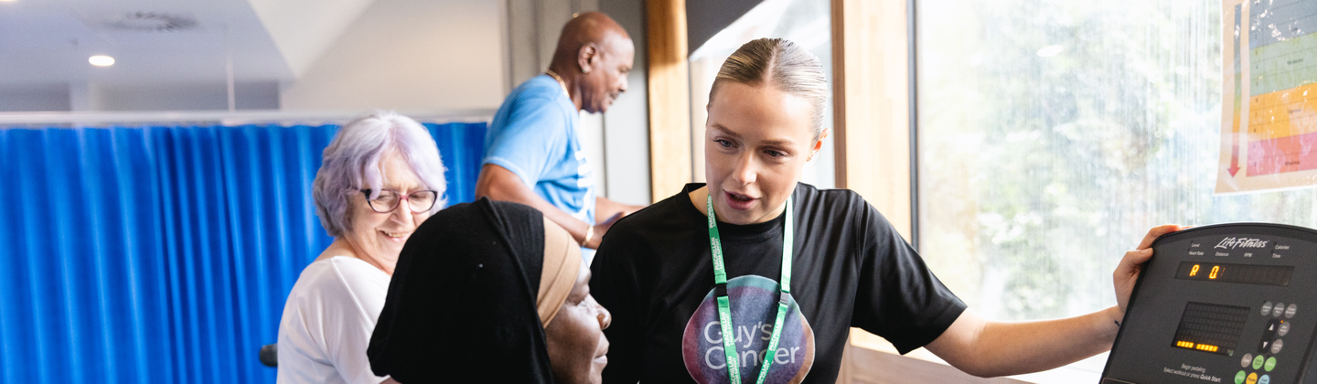 Physiotherapist Isla is standing next to an exercise bike. A patient is using the machine. Behind Isla are other patients using different exercise machines.