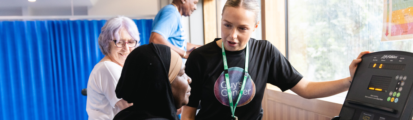 Physiotherapist Isla is standing next to an exercise bike. A patient is using the machine. Behind Isla are other patients using different exercise machines.