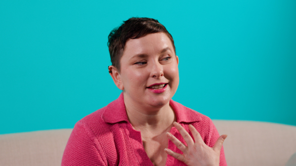 A woman with short hair and pink lipstick is sitting on a sofa telling her story 