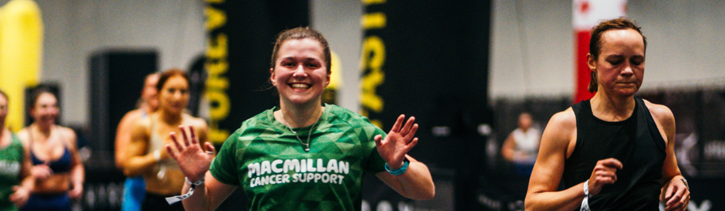 A person wearing a green Macmillan sports top is waving with both their hands. They have their brown hair tied back. They appear to be competing in a HYROX event.