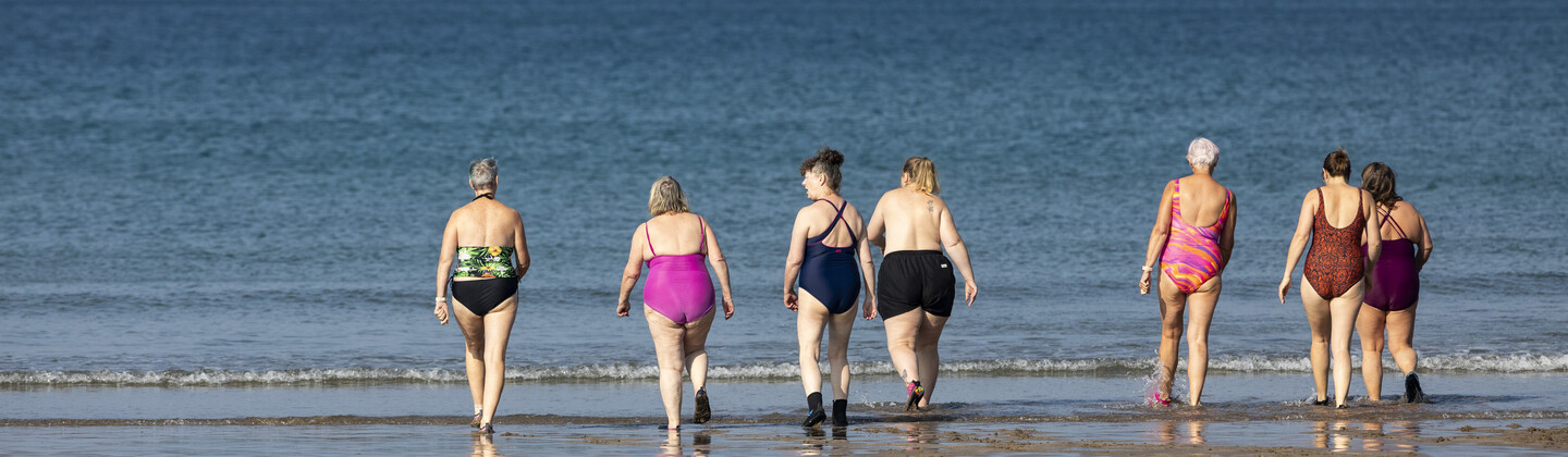 A group of swimmers are walking on the beach towards the sea. They are wearing different swimming suits and gear.