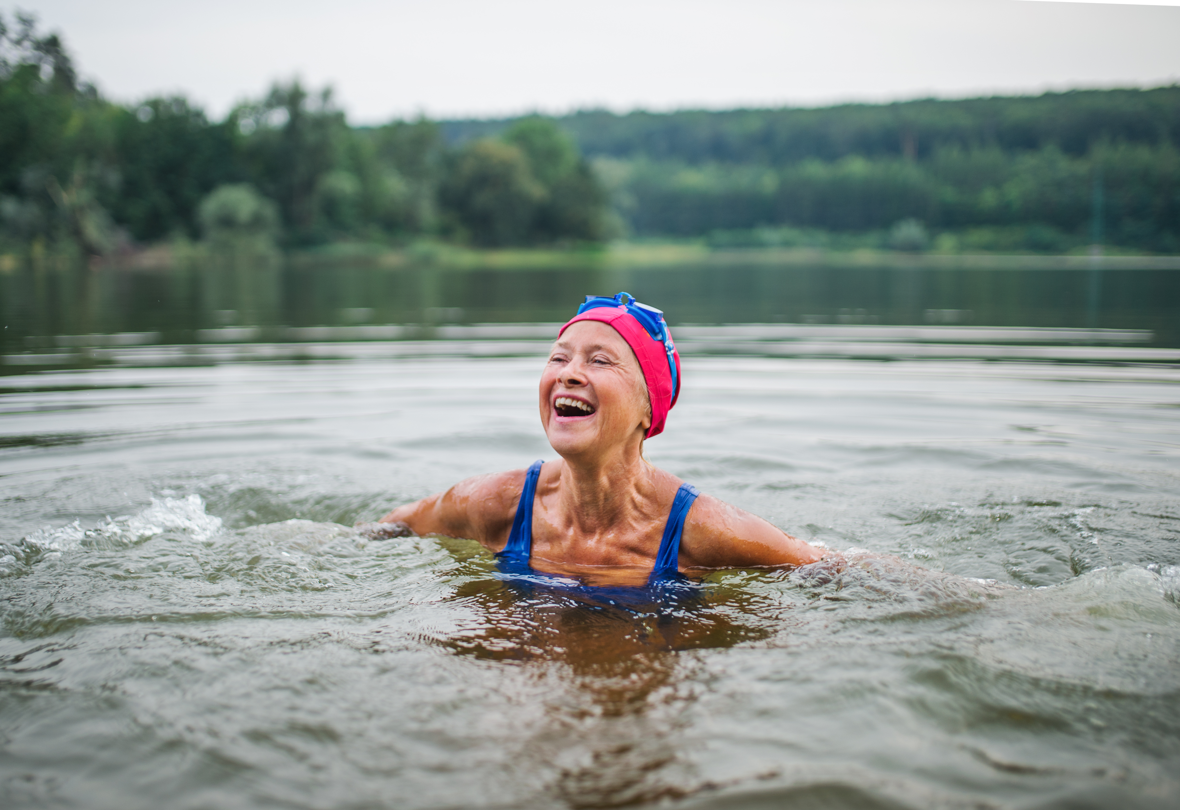An older person is swimming in a lake. They are wearing a pink swimming cap, blue swimming goggles and a blue swim suit. 