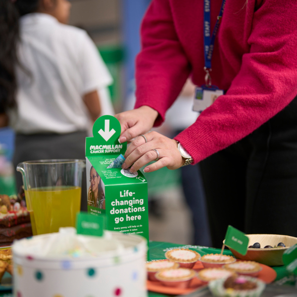 A person in a bright pink jumper is leaning over a table and placing a pound note in a Macmillan collection box. The box is on a table that has been decorated for a Coffee Morning. There are drinks and different sweat and savoury foods on the table. 