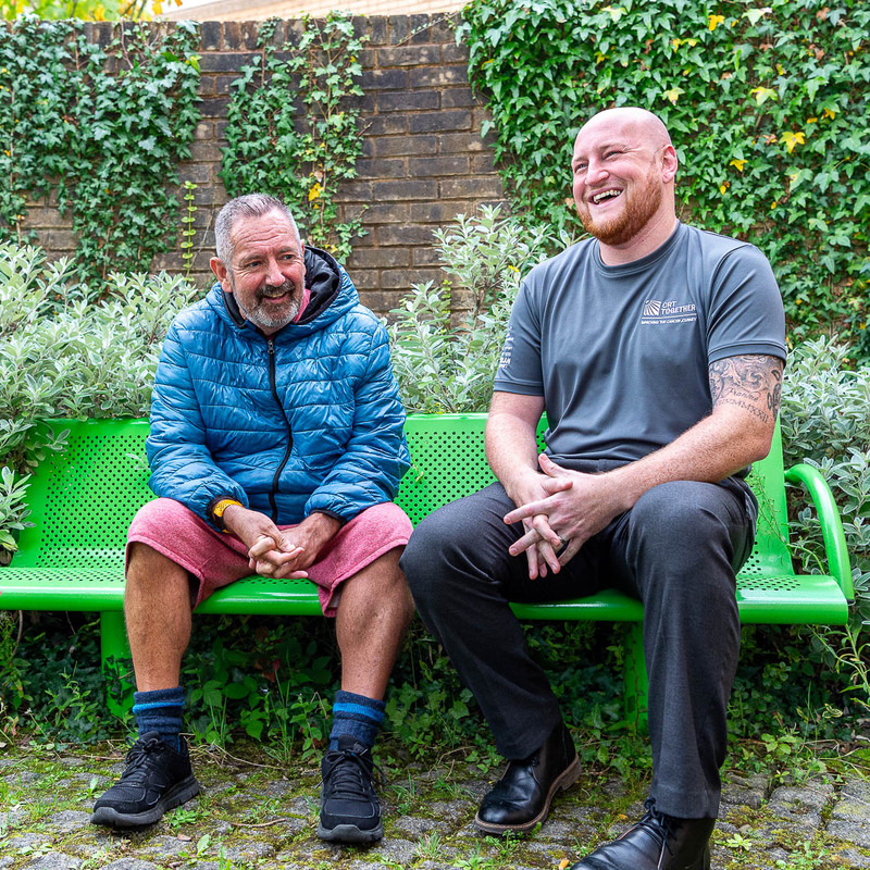 A link worker and a client from CRT Together are sitting on a bench outside laughing together  
