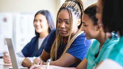 A group of nurses are in a class. Two nurses are looking at a laptop. They are all wearing different coloured scrubs.