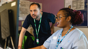 Anthony and another healthcare professional are looking at a desktop computer. They are both wearing NHS healthcare uniforms. They appear to be in a hospital.