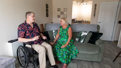 Macmillan Social Care Coordinator Jan Bolton is wearing a bright green patterned dress and is sitting on a sofa. Next to her is a person. They are using a wheelchair. They are inside and talking to each other.
