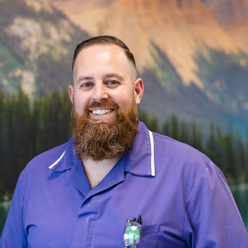 John is a Macmillan Cancer Clinical Nurse Specialist. He has short brown hair and a long brown beard and moustache. He is wearing a blue NHS uniform and standing in front of a landscape back drop.