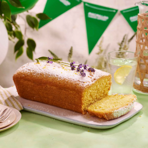 A golden lemon drizzle loaf cake topped with powdered sugar and sprigs of fresh lavender, resting on a white rectangular plate. A slice has been cut to reveal its soft, fluffy interior.