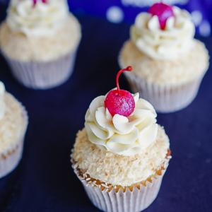 A golden-brown pineapple upside-down cupcake topped with creamy white frosting, a sprinkle of shredded coconut, and a bright red maraschino cherry with its stem.