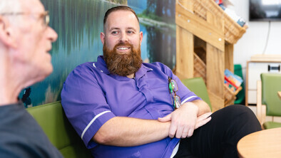 John is sitting down on a green sofa. He is listening to a person speaking. John has short brown hair and a brown beard. John is wearing a purple NHS uniform.