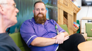 John is sitting down on a green sofa. He is listening to a person speaking. John has short brown hair and a brown beard. John is wearing a purple NHS uniform.