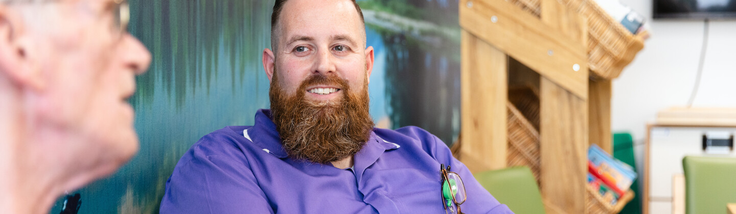 John is sitting down on a green sofa. He is listening to a person speaking. John has short brown hair and a brown beard. John is wearing a purple NHS uniform.