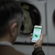 A person is holding out their mobile phone. On the phone is the Online Community homepage. They appear to be in a laundry mat as there are washing machines in the background.