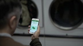 A person is holding out their mobile phone. On the phone is the Online Community homepage. They appear to be in a laundry mat as there are washing machines in the background.