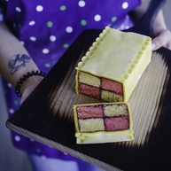 A Battenberg cake with a checkered pattern of pink and yellow sponge squares, bound with apricot jam and wrapped in smooth marzipan with a scalloped top edge. Presented on a wooden tray with one slice cut to reveal its vibrant interior.