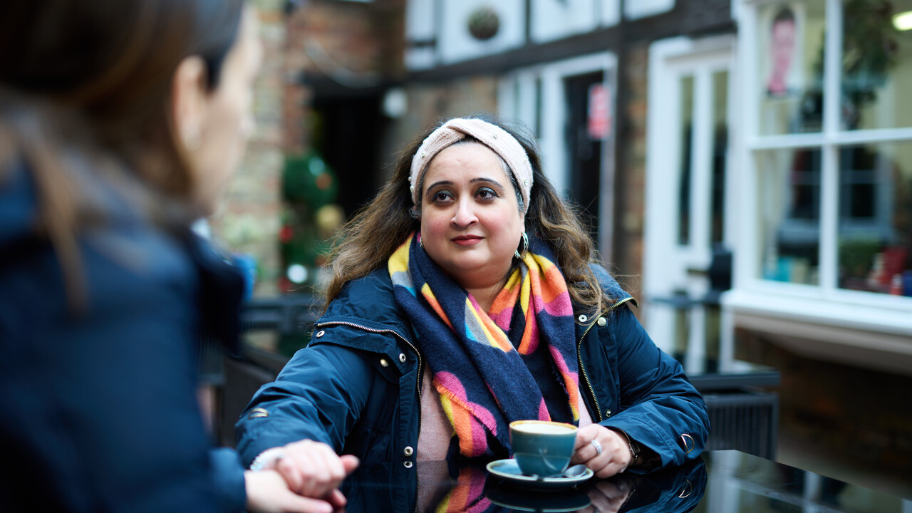 Two people are sitting at a table drinking coffee. One person is holding the other person's hand and looking at them supportively.