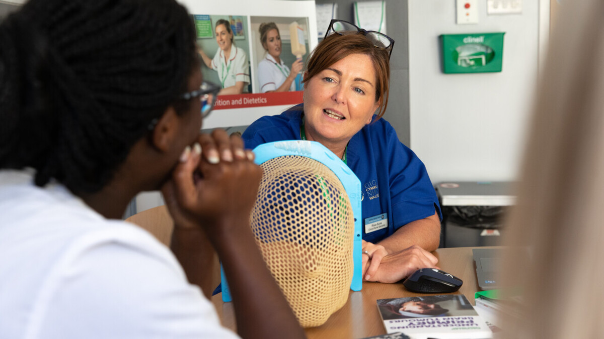 Two people are sitting at a table in a clinic. One is wearing a healthcare professional uniform. There is a radiotherapy mask on the table between them.