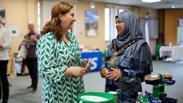 Two women in conversation, one has brown hair and is wearing a green patterned dress. The other is wearing a blue hijab and floral dress, she's also wearing a ceremonial gold chain around her neck
