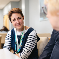 Macmillan Principal speech and language therapist Heulwen Sheldrick is sitting with a group of people. She has short brown hair and is wearing a striped top and blue frock. They appear to be inside a support centre.