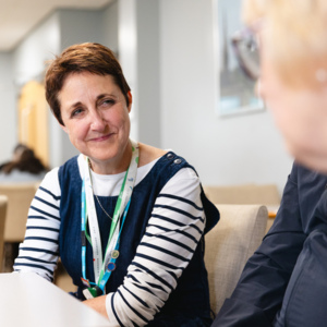 Macmillan Principal speech and language therapist Heulwen Sheldrick is sitting with a group of people. She has short brown hair and is wearing a striped top and blue frock. They appear to be inside a support centre.