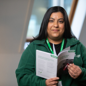 A Macmillan money adviser is holding open a Macmillan booklet. They have long dark hair and are wearing a Macmillan branded green jacket. 