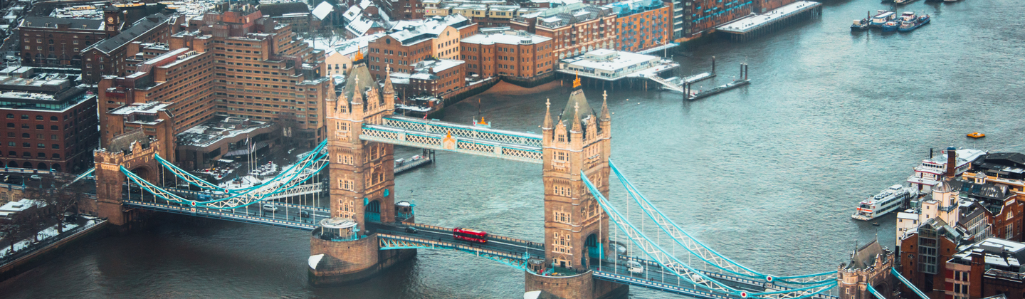 An aerial view of Tower Bridge in London. There is snow on the ground and on different buildings near the bridge. 
