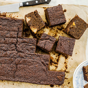A rich, dark brown pear and ginger cake with a cracked surface sits on parchment paper, its moist texture revealed by square slices showing chunks of pear nestled in spiced ginger batter.