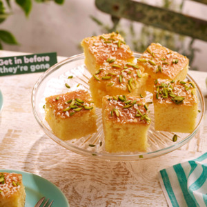 Six golden brown squares of moist revani cake on a glass plate and topped with shredded coconut and finely chopped pistachios.