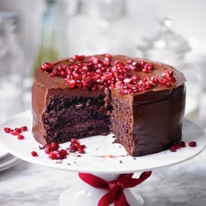 A rich chocolate cake with glossy chocolate frosting, topped generously with vibrant red pomegranate seeds. Displayed on a white cake stand with a large slice cut out, the cake reveals its moist, dense interior.