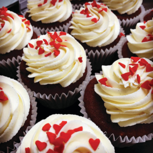 Red velvet cupcakes topped with generous swirls of creamy white frosting and decorated with red heart-shaped sprinkles. The rich red sponge peeks through beneath the frosting.