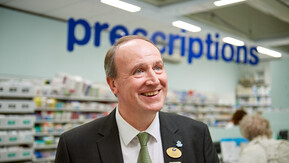 Graham Pickup is a Boots Macmillan Information Pharmacist. He is wearing a suit with a black coat and green patterned tie. Graham is smiling and has short brown hair and blue eyes. Behind him is a pharmacy with the sign prescriptions in large blue print. 