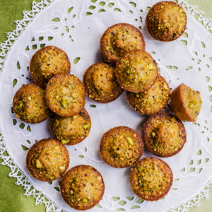 A batch of freshly baked pistachio mini muffins with golden-brown crusts and crunchy green pistachio pieces, arranged on a white lace doily. Set against a green tablecloth, the close-up view makes these nutty treats look irresistibly warm and inviting.