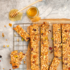 A batch of freshly baked granola bars packed with oats, nuts, seeds, and dried fruits rests on a cooling rack. Rectangular pieces are cut and ready to serve, while a jar of golden honey with a dripping dipper adds a glossy, indulgent touch.