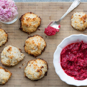 Seven golden-brown coconut macaroons with toasted edges rest on a wooden board, paired with a bowl of vibrant raspberry jam and a spoonful ready to serve.