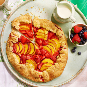 A golden-brown rustic fruit galette brimming with juicy peach and strawberry slices sits on a light blue wooden board. It's surrounded by fresh berries, steaming cups of tea or coffee, and a pitcher of cream.