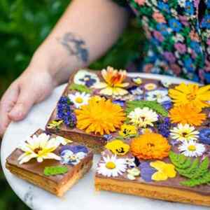 A beautiful millionaire's shortbread. Sticky caramel, smooth chocolate and vibrant edible flowers—orange marigolds, white daisies, purple lavender, and yellow pansies. A slice has been cut to reveal luscious chocolate layers and a biscuit base.
