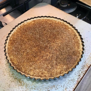 A custard tart in a metal tart tin is on top of a white counter top. The tart is topped with sprinkled grated nutmeg. 