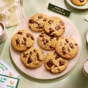 A plate of golden-brown chocolate chip cookies with gooey melted chunks sits on a light green wooden table. Two frothy coffees in white and beige mugs, partially eaten cookies on side plates, and green-striped napkins create a cozy, inviting scene.