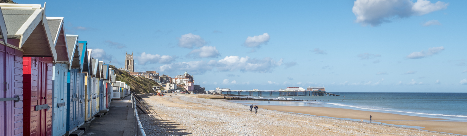 Beach huts in a line on the left against a large beach and the sea on the right. A pier can be seen in the distance.